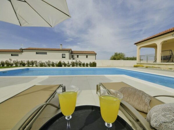 A poolside view with two glasses of yellow drink on a tray, a lounge chair, and a building with a tiled roof in the background. - Vacation home Nisa by Interhome (Ražanac)