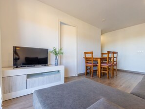 Ein modernes Wohnzimmer mit einem Flachbildfernseher auf einer weißen Konsole, einem Esstisch mit vier Holzstühlen und einem grauen Sofa.