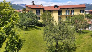 A yellow building with a red roof, surrounded by greenery and mountains in the background. - Lovely apartment for 3 people with WIFI, TV and patio (Germignaga)