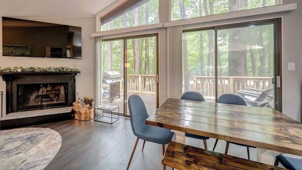 A spacious living room with a large wooden dining table, a fireplace, and large windows offering a view of the outdoors. - Cozy Pet-Friendly w Fire Pit, Wifi & Pool Access (Hedgesville)