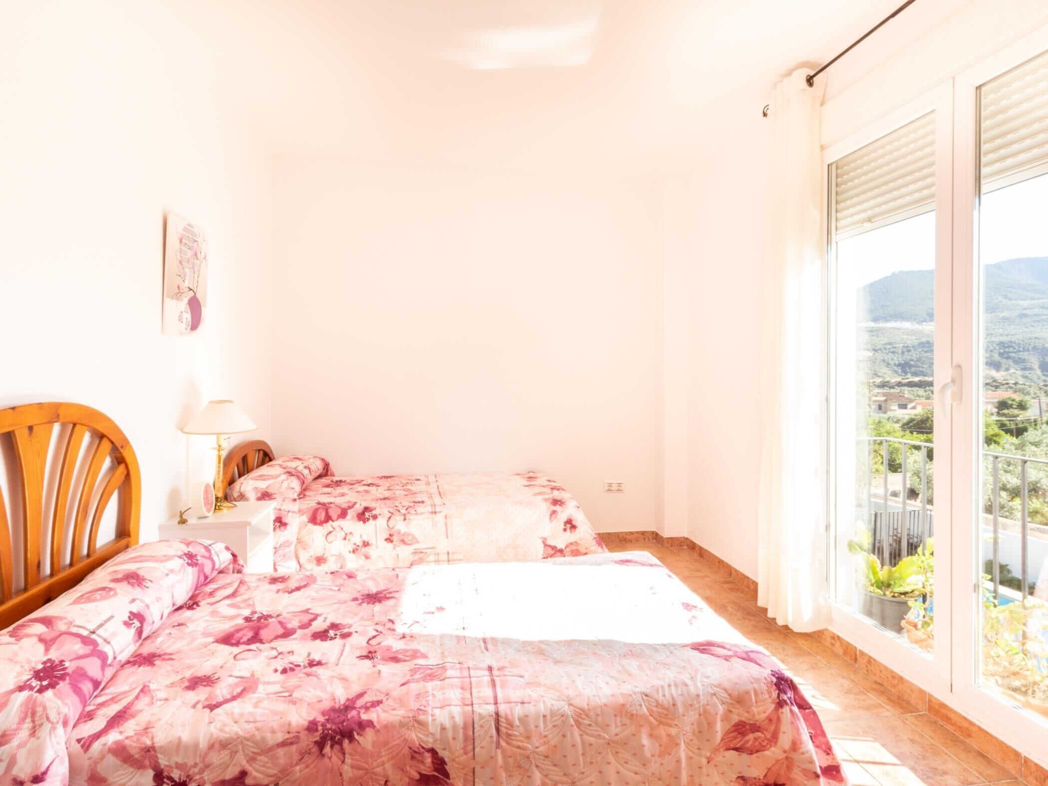 A bedroom with two beds, a wooden chair, a bedside table with a lamp, and a large window offering a view of a mountainous landscape.