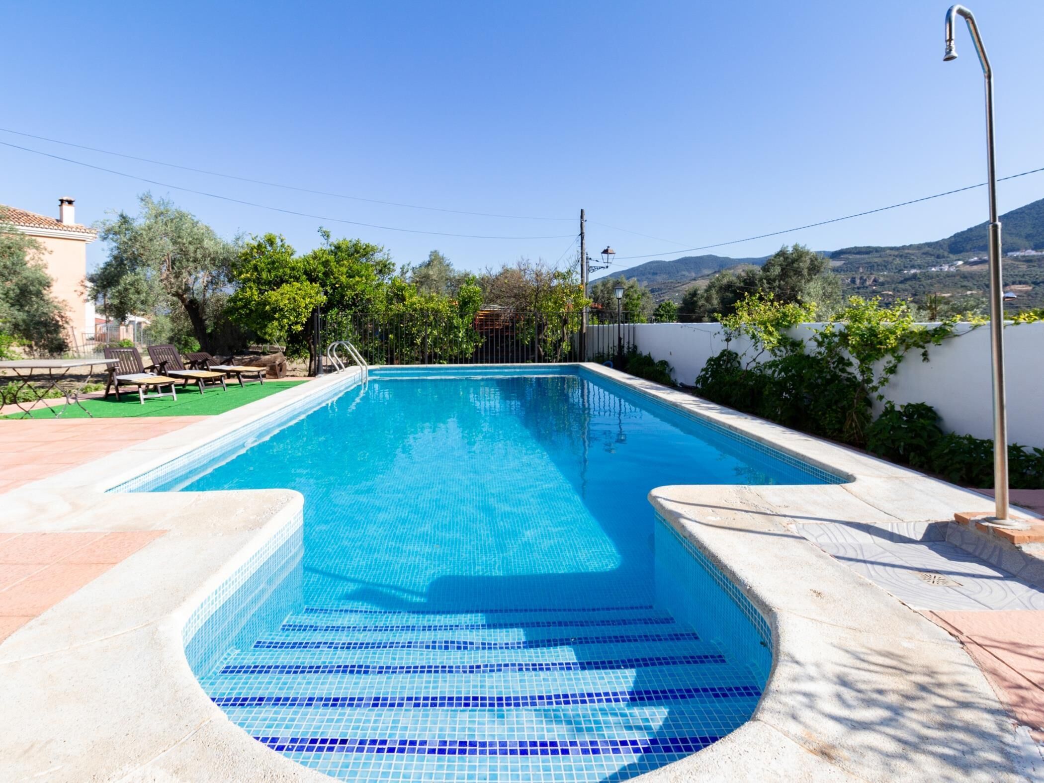 A rectangular swimming pool with clear blue water, surrounded by a white wall and a metal fence. There are lounge chairs and a table on the left side, and a lamp post on the right.