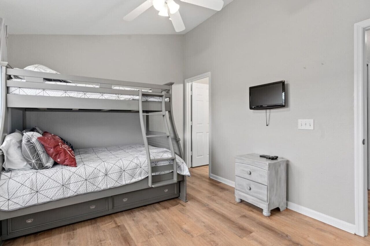 Una cama litera con escalera, una mesita de noche pequeña, un televisor en la pared y una puerta que da a otra habitación.