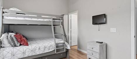 Una cama litera con escalera, una mesita de noche pequeña, un televisor en la pared y una puerta que da a otra habitación.