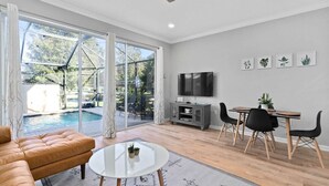 Een ruime woonkamer met een grote schuifpui naar een zwembad, een comfortabele bank, een eettafel met stoelen en een flatscreen televisie aan de muur.