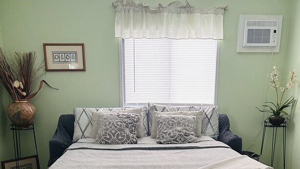 A bedroom with a bed, a window with blinds, a potted plant, and a wall-mounted air conditioner. - Puakenikeni Hilo Hale (Hilo)