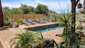 Un espace piscine agrémenté d’une terrasse en bois, de chaises longues et de plantes tropicales.