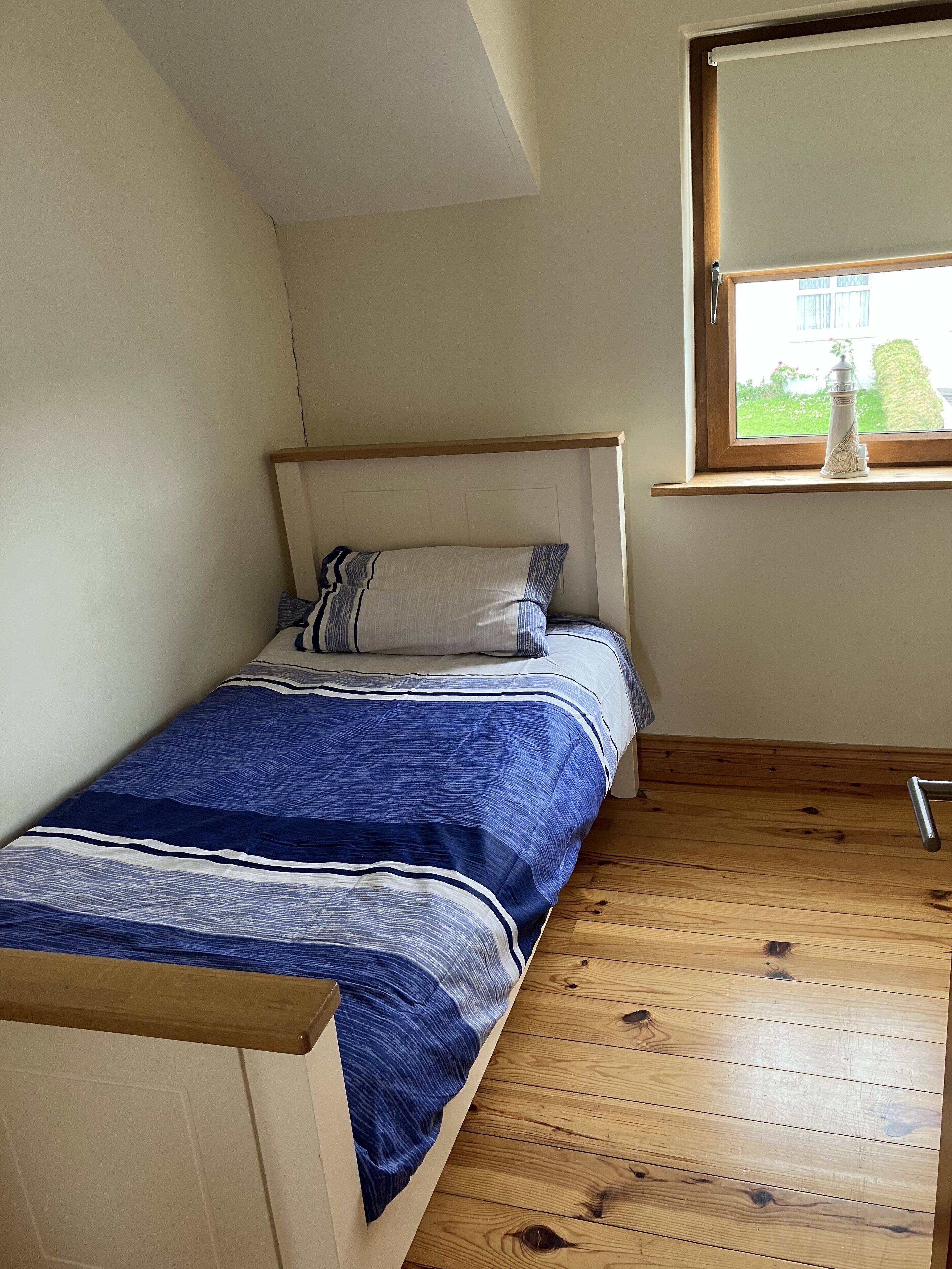 A small, single-bed room with a wooden floor, a window with a view of greenery, and a bedside table with a lamp.