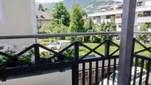 Een balkon met een zwart metalen hekwerk, betegelde vloer en uitzicht op een groen landschap met gebouwen en auto's op de achtergrond.