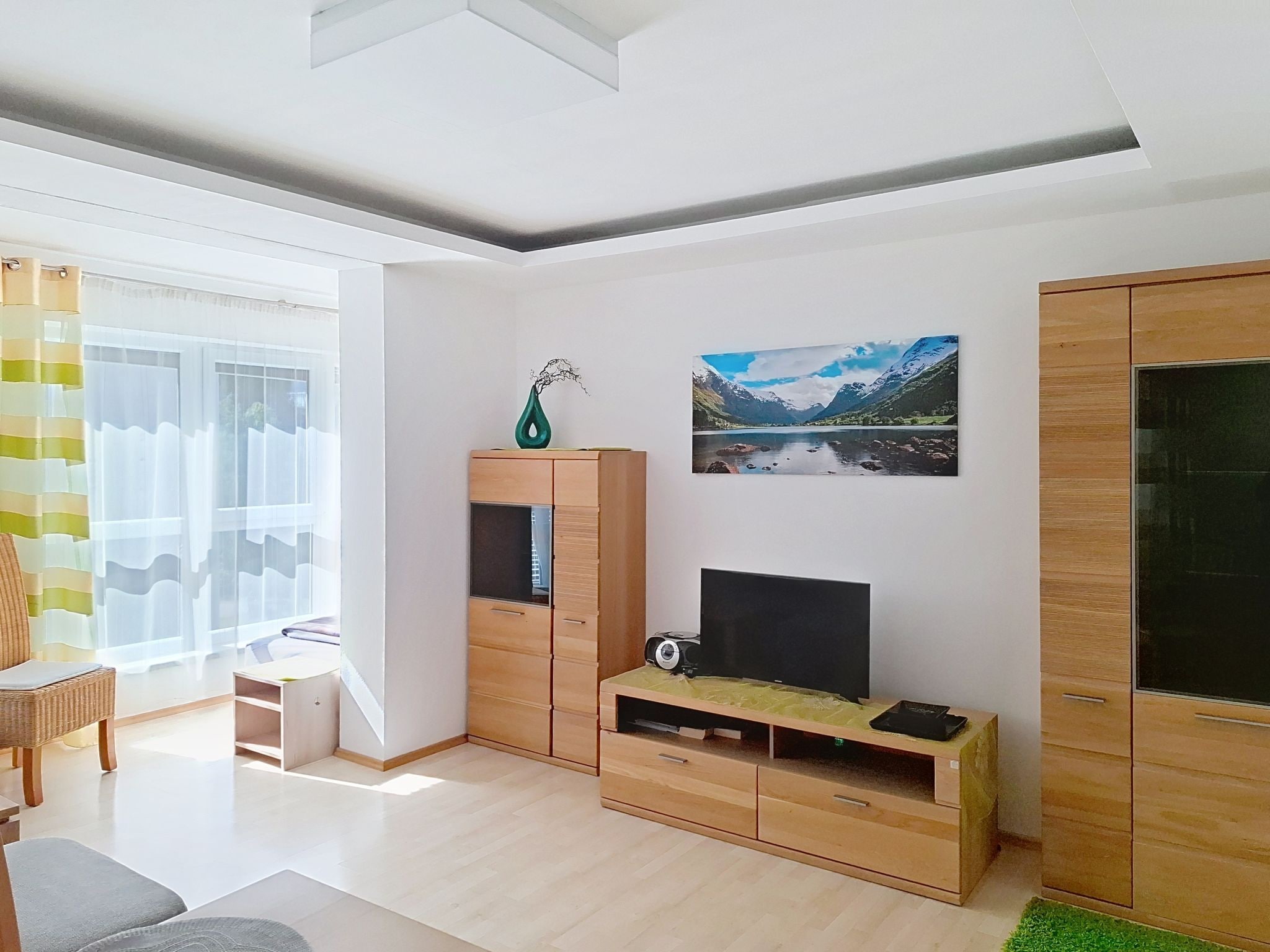 A modern living room with a flat-screen TV, wooden cabinets, and a large window with curtains.