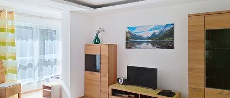 A modern living room with a flat-screen TV, wooden cabinets, and a large window with curtains.