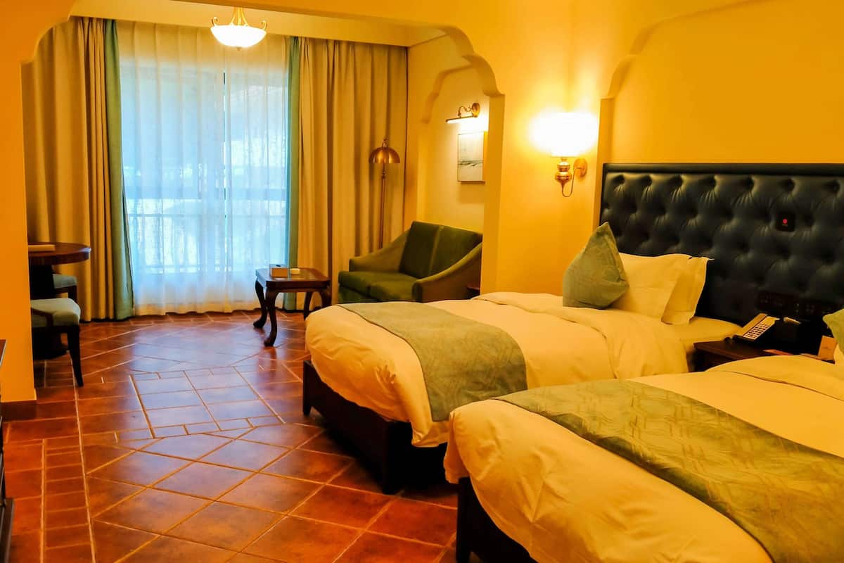 Une chambre d’hôtel avec deux lits, une chaise, une petite table et une vue sur l’extérieur par une fenêtre avec des rideaux.