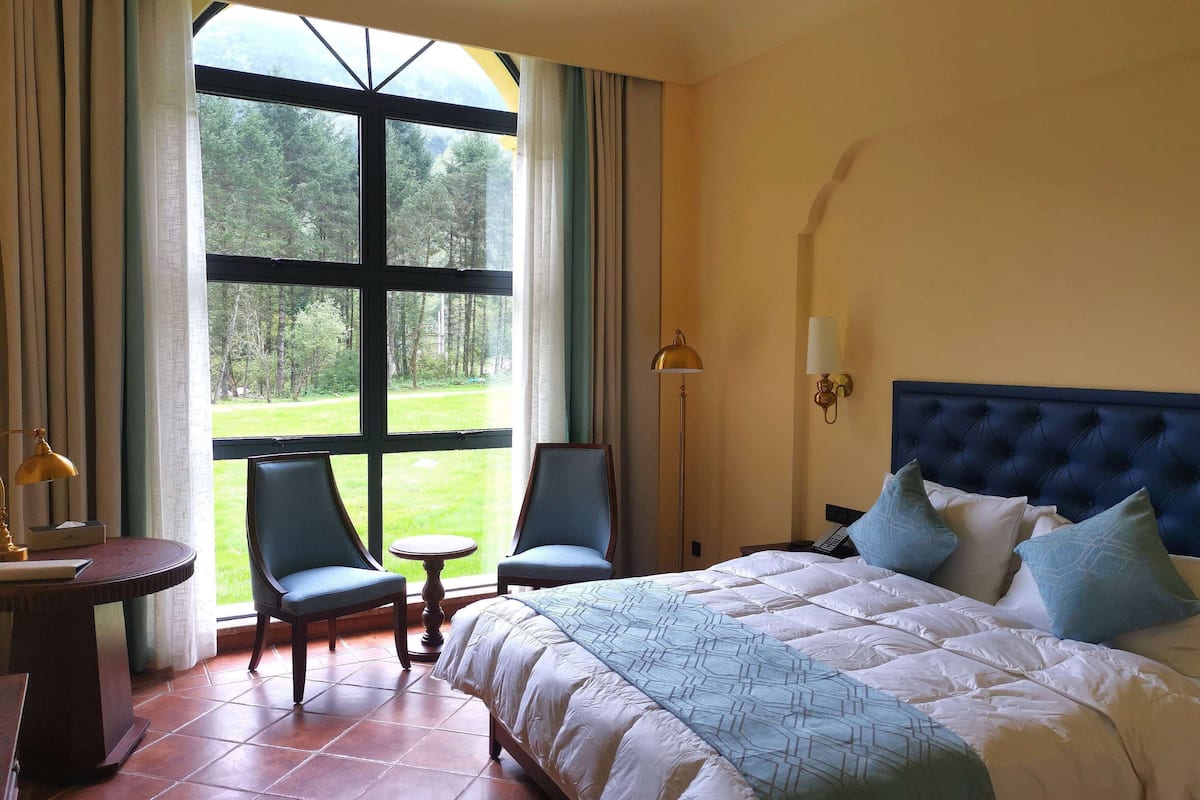 Une chambre avec une grande fenêtre, un lit avec une literie bleue et blanche, deux chaises, une petite table et une lampe.
