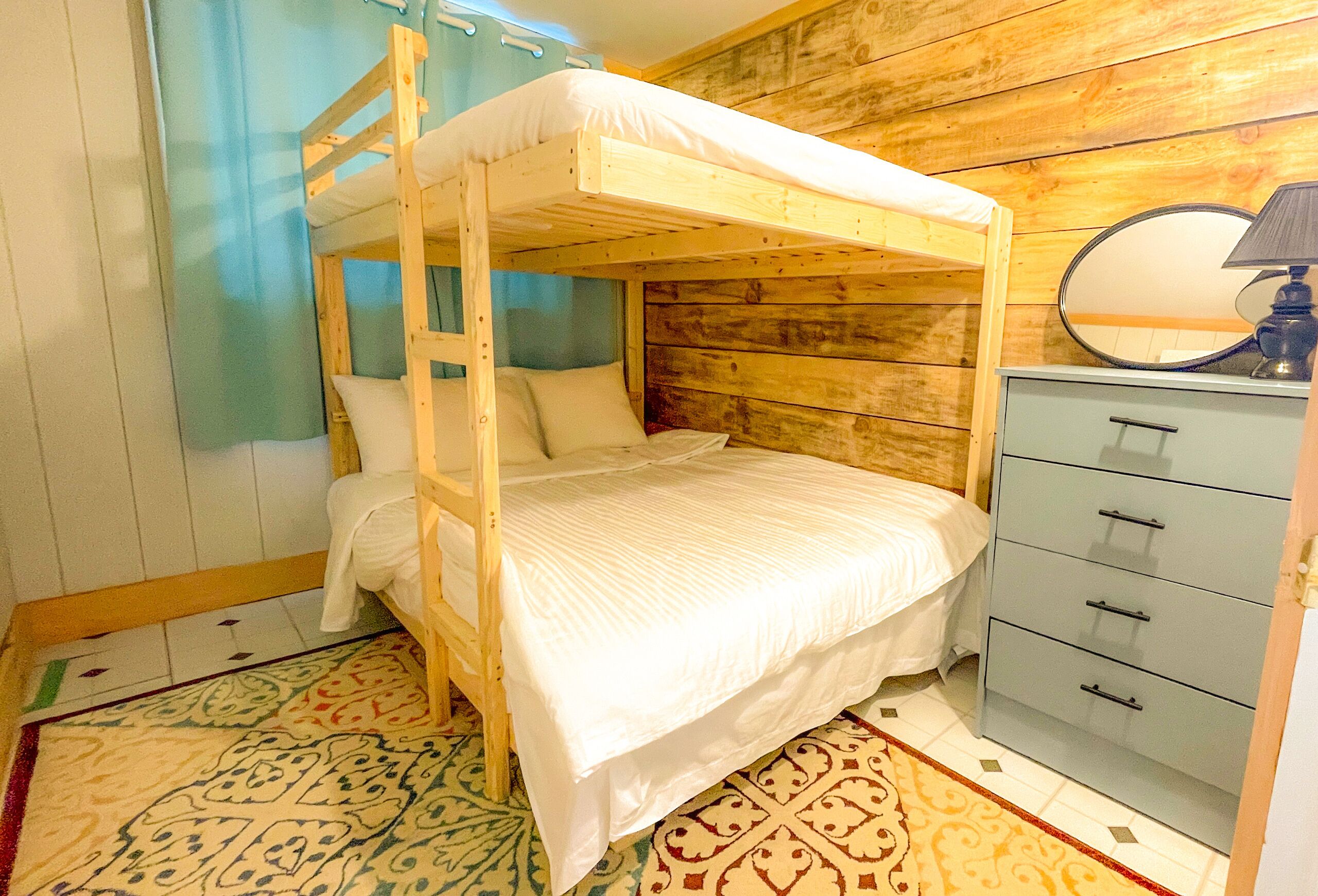 A bunk bed with a wooden frame and white bedding, a small dresser with drawers, and a patterned rug on the floor.