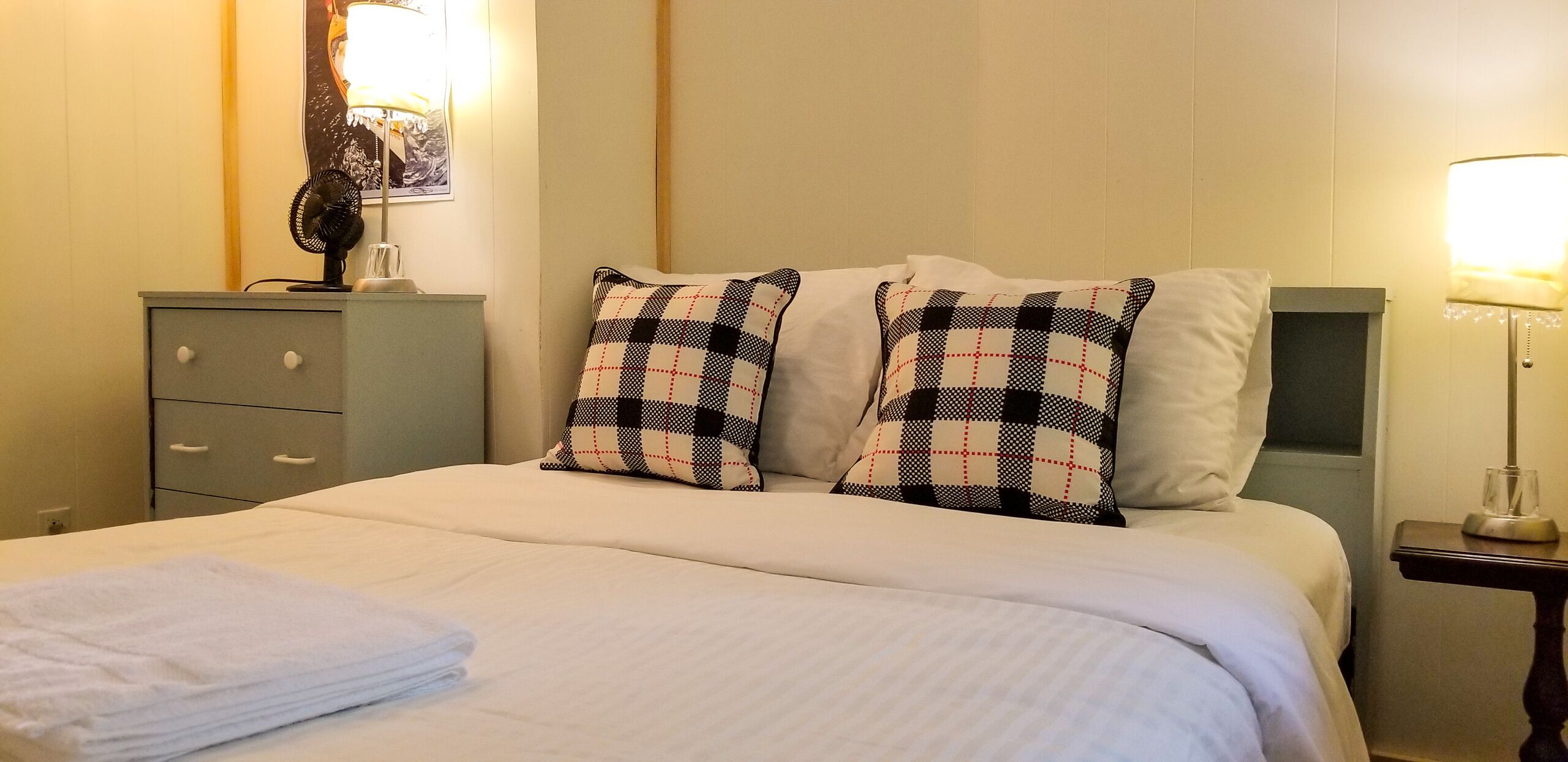 A neatly made bed with checkered pillows, a bedside table with a lamp, and a small cabinet with drawers.