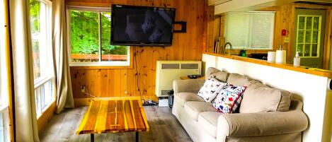 Un salon chaleureux avec un plancher en bois, un canapé beige, une table basse en bois, un téléviseur à écran plat fixé au mur et une fenêtre donnant sur un paysage verdoyant.