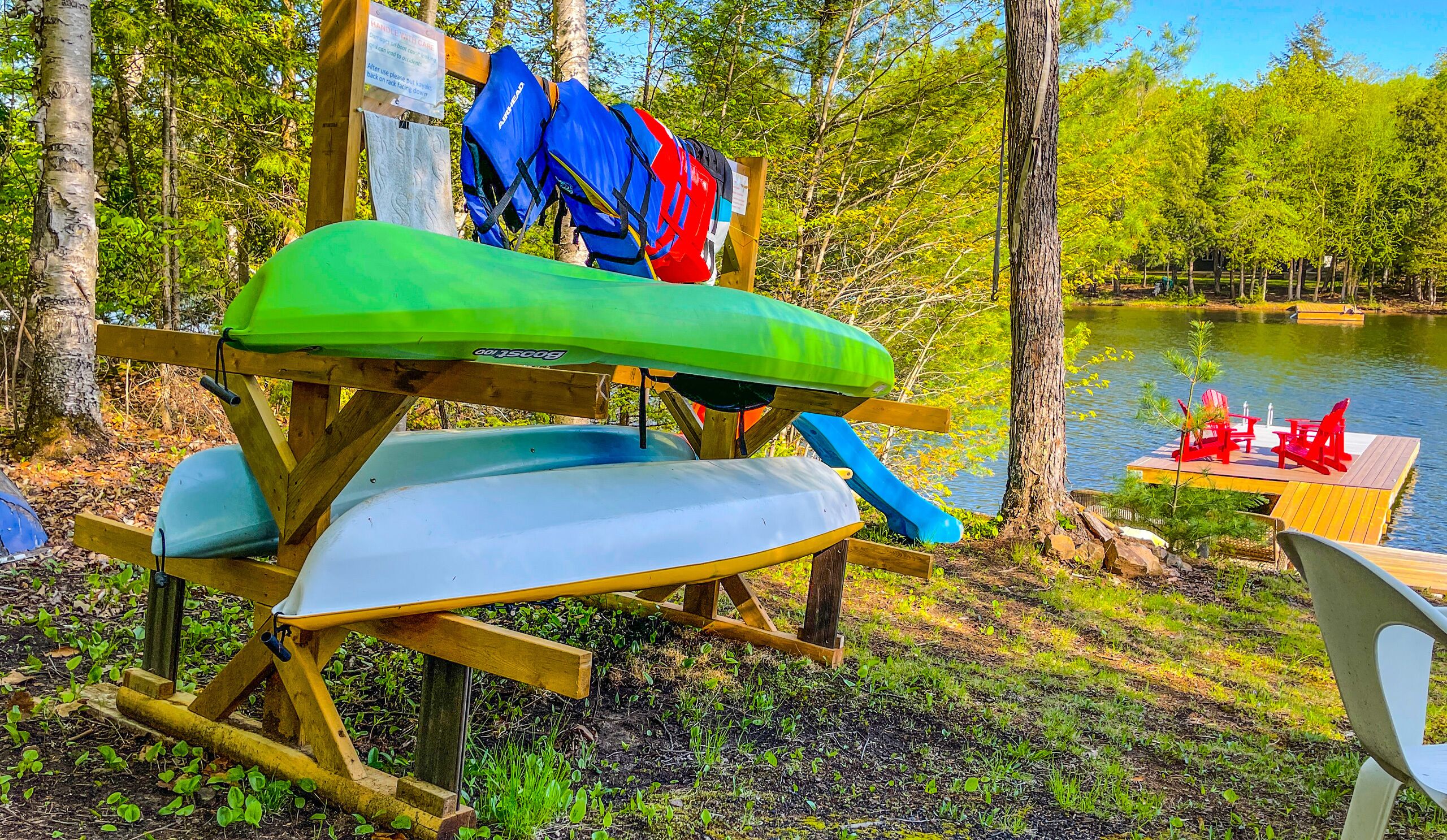 A kayak rack with a green kayak and a blue kayak, a wooden deck with a dock, and a lake with red chairs and a table.
