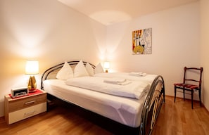 Una cama bien hecha con almohadas, una mesita de noche con una lámpara, y una silla en una habitación con suelo de madera y un cuadro en la pared.