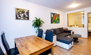 Un salon moderne avec une table à manger en bois, deux canapés, une table basse et deux fauteuils. Deux tableaux ornent les murs, et une plante en pot est posée dans un coin.
