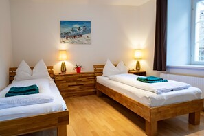 Une chambre d’hôtel avec deux lits en bois, du linge de lit blanc, un tableau représentant un paysage de montagne enneigé et une fenêtre avec des rideaux.