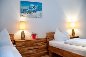 A hotel room with two beds, wooden headboards, bedside lamps, a red potted plant, and a painting of a snowy mountain scene.