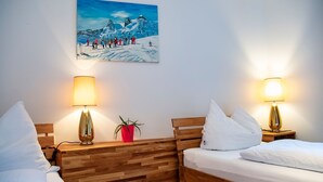 A hotel room with two beds, wooden headboards, bedside lamps, a red potted plant, and a painting of a snowy mountain scene.