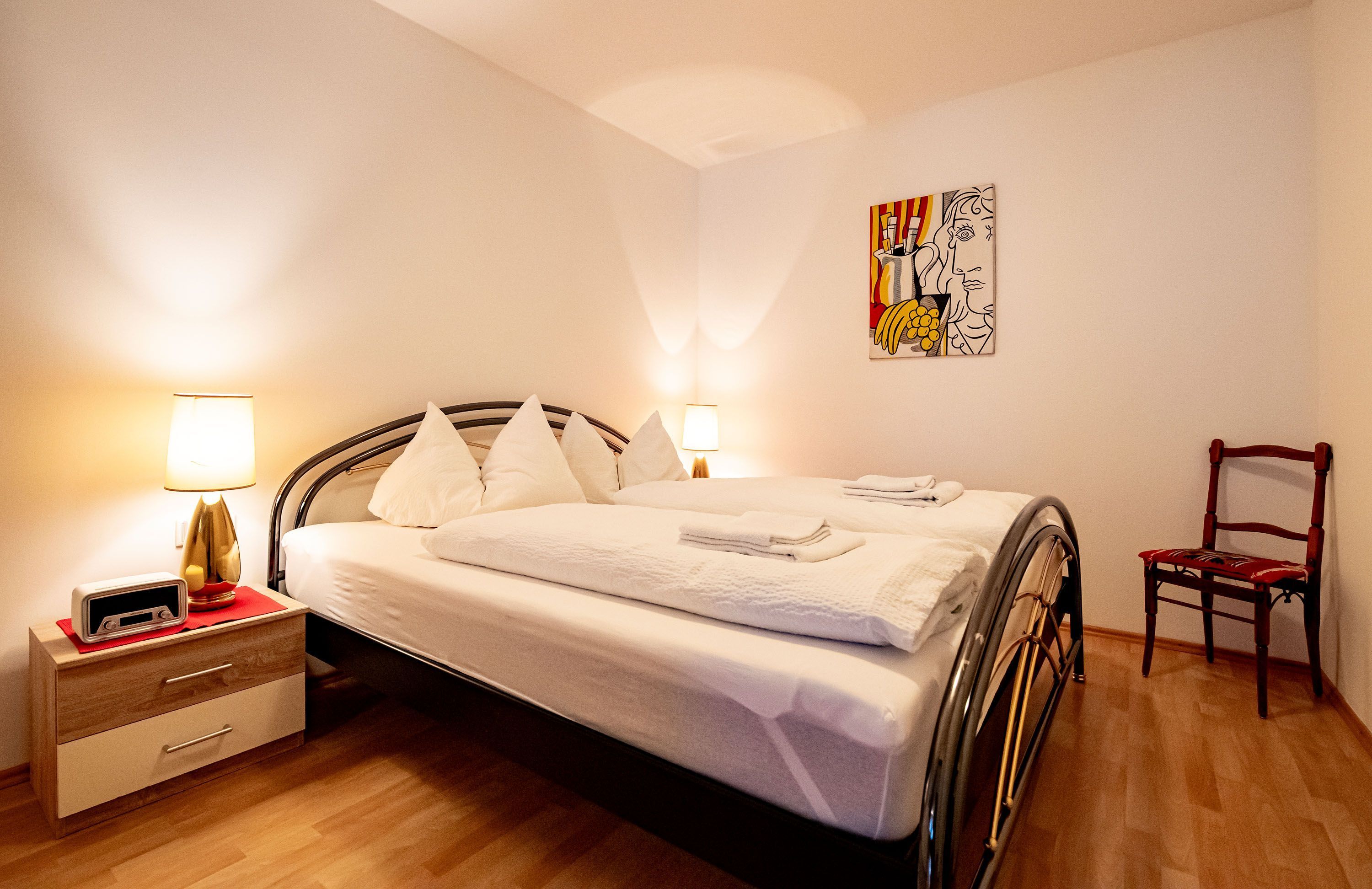 A neatly made bed with pillows, a bedside table with a lamp, and a chair in a room with wooden flooring and a painting on the wall.