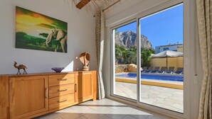 A room with a wooden cabinet, a painting of an elephant, and a view of a pool and mountains through large sliding glass doors. - El Elefante - Javea, Costa Blanca (Javea)