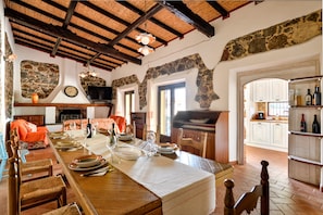 A dining area with a long wooden table set for a meal, stone walls, a stone fireplace, and a kitchen visible in the background. - Country House by the Lake with Heated Private Pool, Lake & Mountain Views, Garden (Dorgali)