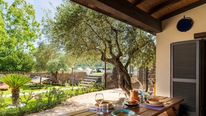 Taula de fusta a l'aire lliure, coberta amb plats, gots i una gerra, sota una zona protegida amb un arbre i vista a un jardí i unes escales.