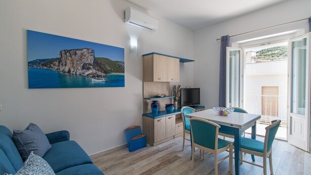 A room with a sofa, a dining table with chairs, a TV, and a painting of a coastal scene. - Apartment Blu with Mountain View, Balcony, A/C & Wi-Fi (DORGALI)