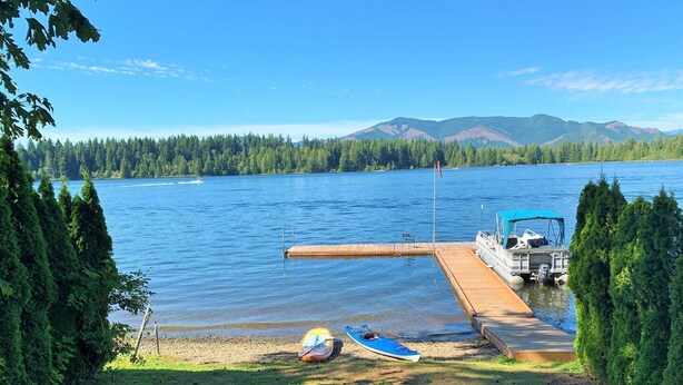 Un muelle junto al lago con un bote y un kayak, rodeado de árboles y montañas al fondo.