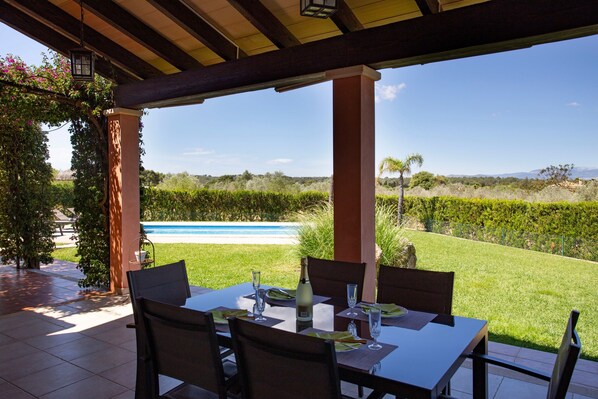 Un porche cubierto con una mesa lista para comer, con vista a una piscina y jardín ajardinado.