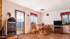 A cozy living room with a wood-burning stove, a dining area with wooden chairs, a television, and a large window with curtains. - Apartment "Baita Località Cap" with Garden & Mountain View (Roncone)