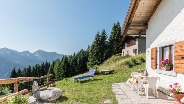 A patio with a stone table, chairs, a blue lounge chair, and a view of mountains. - Apartment "Baita Località Cap" with Garden & Mountain View (Roncone)