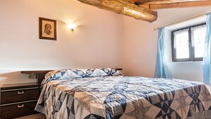 A bedroom with a bed, a nightstand, a wall-mounted light, a window with curtains, and a wooden ceiling beam. - Apartment "Baita Località Cap" with Garden & Mountain View (Roncone)