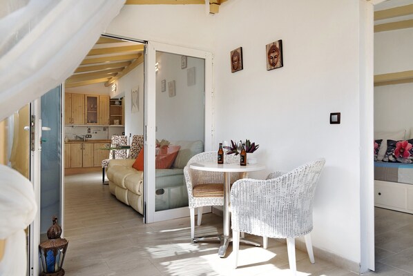 Una habitación con una mesa de comedor de mimbre blanca, dos sillas de mimbre, un sofá y una puerta de cristal que da a una cocina.
