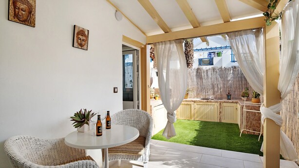En terrasse med fletningsmøbler, et lille bord og udsigt til en baghave med grøn græsplæne og træhegn.