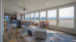 A spacious living area with a large window overlooking a beach, a ceiling fan, and a dining area with a view.