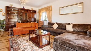 A living room with a sofa, armchair, and a coffee table. There is a large wooden cabinet with glass doors and a chandelier.