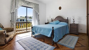 A bedroom with a bed, a chair, a nightstand, a dresser, and a balcony with a railing.