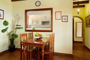 Un coin repas avec une table en bois, des chaises, un vase avec des plantes, un panier de fruits et une cuisine visible à travers une fenêtre.