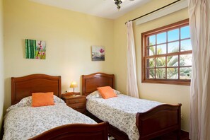 Une chambre avec deux lits en bois, une table de chevet avec une lampe, une fenêtre avec des rideaux et des œuvres d’art accrochées au mur.