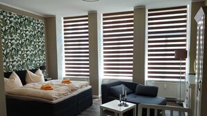 A modern hotel room with a large bed, a sofa, a small table, and a view through large windows with blinds.