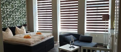 A modern hotel room with a large bed, a sofa, a small table, and a view through large windows with blinds.