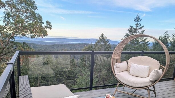 A balcony with a wicker chair and a cushioned bench, overlooking a forested landscape with mountains in the distance. - Entire room. Ocean,mountain view, hot tub,garden,deck,next to Mount Maxwell park (Salt Spring Island)
