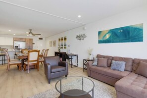 A modern living room with a brown sofa, a glass coffee table, and a dining area with wooden chairs and a white table. - New Photos! Direct beach access+pool+parking (San Diego)