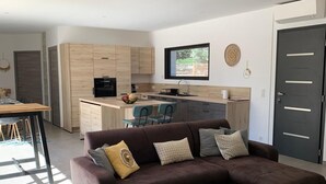 A modern living room with a sectional sofa, a small round table, and a yellow armchair. A kitchen with wooden cabinets and a dining area with a table and chairs is visible in the background. - Villa Pienezza 8p 4 bedrooms very quiet Pinarellu 5min from beaches Heated swimming pool (Pinarellu)