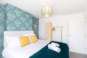 Hotellihuone, jossa on sänky, valkoiset vuodevaatteet, turkoosinsävyinen korosteseinä, puuovi ja riippuvalaisin.