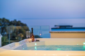 A hot tub with a glass barrier, candles, and bottles, overlooking a body of water and a distant boat. - Penthouse Apartment Perla with Jacuzzi (Novalja)